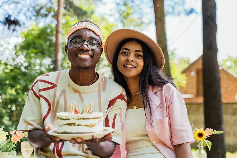 A smiling young man in glasses and a woman in a pink shirt and sun hat holding a layered cake with lit candles outdoors under tall trees