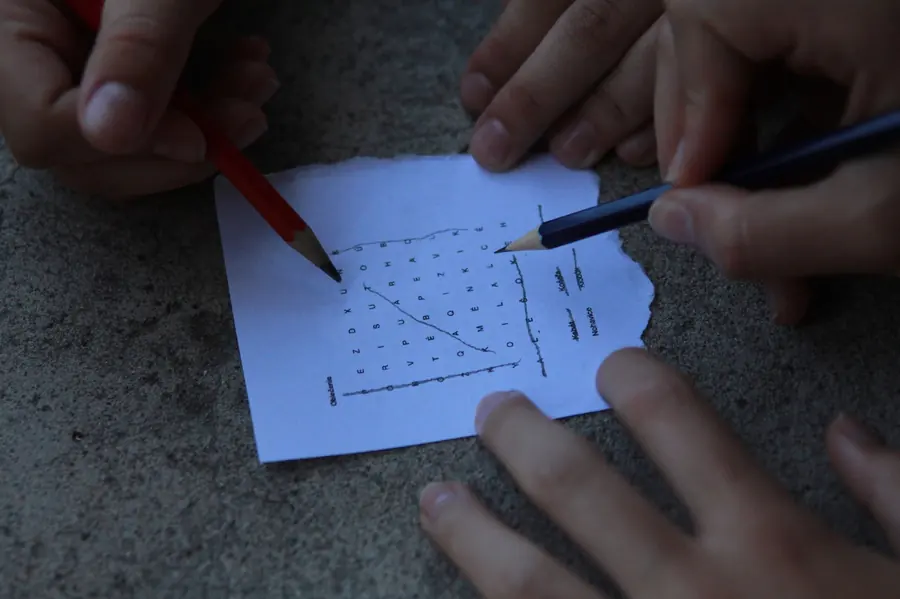 Three people’s hands with two pencils working through a word-search puzzle printed on a torn scrap of paper, on a concrete surface