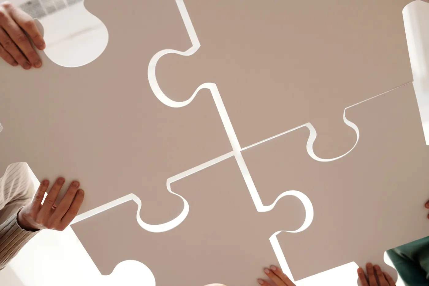 Four pairs of hands lifting oversized white jigsaw pieces so that they lock together, shot from below against a bright ceiling