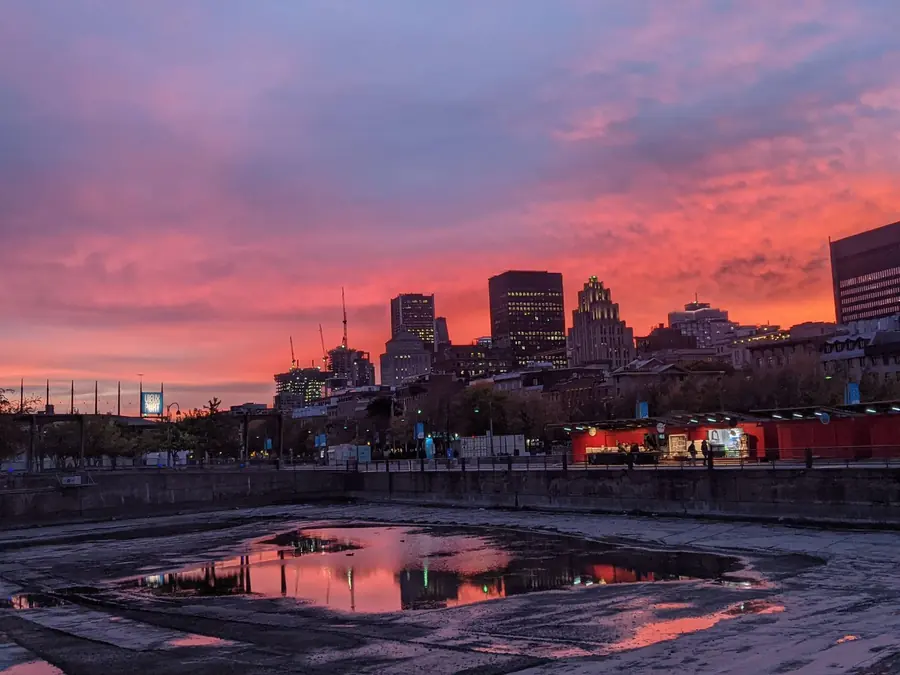 A pink and orange sunset over the Old Port of Montréal, reflected in rainwater pooled on the empty basin in front of the skyline