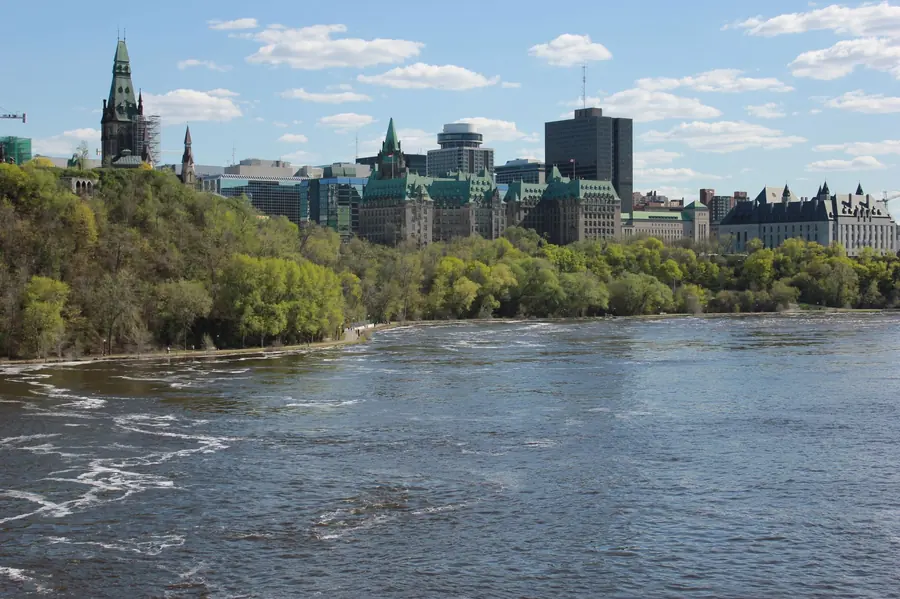 The Parliament Buildings and Supreme Court above a wooded bank of the Ottawa River in spring, with fast water in the foreground